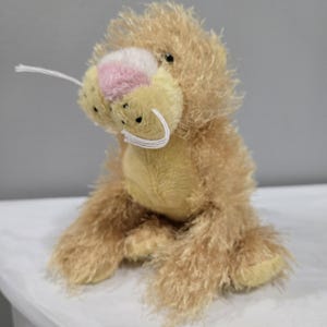 May include: A plush toy lion with a light brown and yellow fur. The lion has a pink nose and white whiskers.