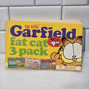 May include: A yellow, green and purple book cover with the title "Garfield Fat Cat 3-Pack" and a cartoon image of Garfield the cat. The book cover also includes a price tag with the text "£20.85 value now only £9.95".
