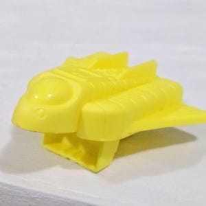 Könnte beinhalten: Ein hellgelbes Spielzeug-Space Shuttle mit abgerundetem Cockpit, Flügeln und rechteckiger Basis. Das Spielzeug besteht aus Kunststoff und hat eine glatte, glänzende Oberfläche. Das Design ist einfach und stilisiert.