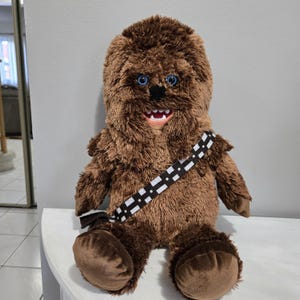 May include: A plush Chewbacca doll with brown fur, blue eyes, and a black nose. The doll has a black and white bandolier across its chest and a smiling mouth. The doll is sitting on a white surface.