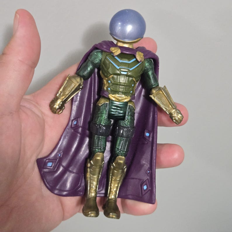 May include: A Mysterio action figure. The figure has a purple cape, a gold and green armored suit, and a clear, spherical helmet. The figure is held in a hand against a gray background. The figure's boots and arm guards are gold.