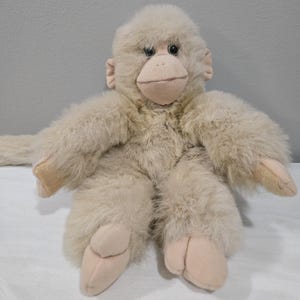 May include: A plush monkey toy in a seated position. The toy is primarily beige with a soft, furry texture. It has a light brown face, ears, hands, and feet. The monkey's arms are outstretched, and it has a long tail.