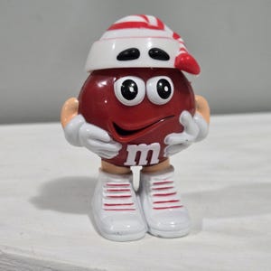 May include: Red M&M character figurine wearing a white and red striped hat and white shoes with red stripes. The character has a big smile and the M&M logo on its chest.