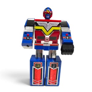 May include: A vintage-style robot action figure featuring a blue, red, and silver colour scheme. The robot has a blocky torso with a yellow V-shaped emblem, a helmeted head, and rectangular legs decorated with red panels and small musical note symbols.