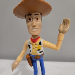 Pu&ograve; includere: Una figurina di plastica di Woody di Toy Story. Indossa il suo cappello da cowboy, il gilet e i jeans caratteristici. Sta salutando con la mano destra.