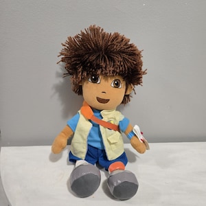 May include: A plush doll featuring a character with brown yarn hair, brown eyes, and a tan complexion. The doll wears a blue short-sleeved shirt, a light yellow vest, blue shorts, and grey shoes. An orange strap crosses the chest, and a blue wristband is visible.