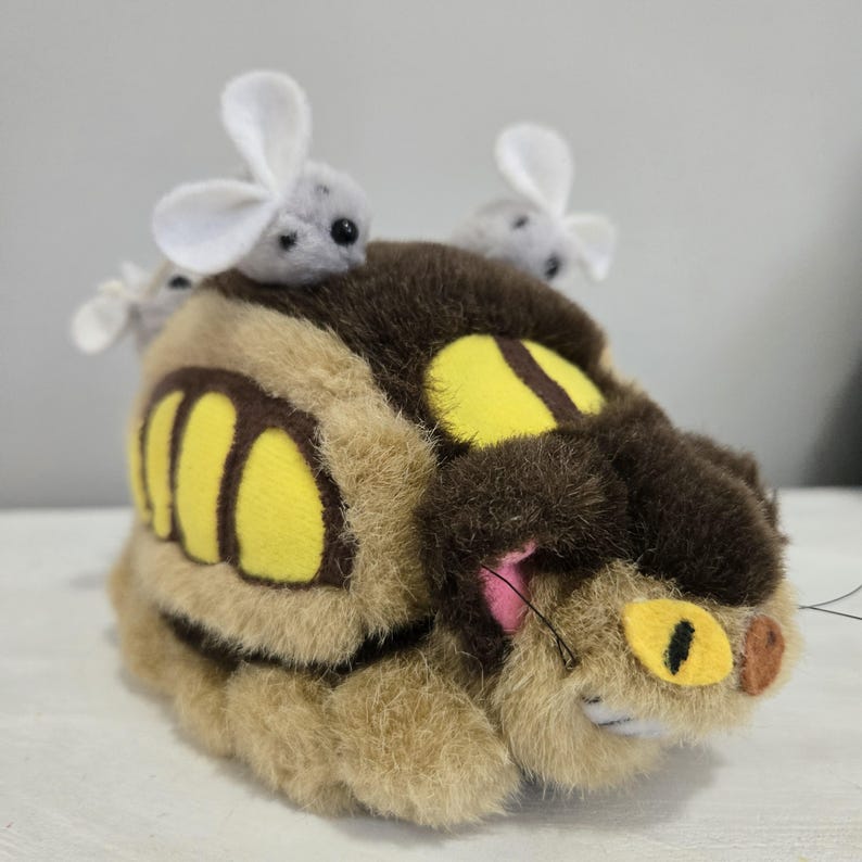 May include: A plush toy resembling a character from a popular animated film. The toy is primarily brown and tan with yellow accents and a dark brown top. Small gray mice with white ears are perched on top of the toy.
