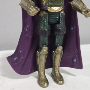 May include: A close-up of a Mysterio action figure. The figure has a purple cape with blue diamond accents, gold boots, and green and gold armor. The figure is standing on a white surface.