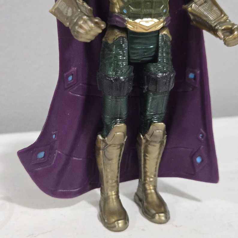 May include: A close-up of a Mysterio action figure. The figure has a purple cape with blue diamond accents, gold boots, and green and gold armor. The figure is standing on a white surface.