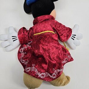 Pu&ograve; includere: Un giocattolo di peluche di Topolino che indossa un cappotto di velluto rosso con note musicali ricamate sul bordo. Il cappotto ha una cintura di corda dorata e il giocattolo ha guanti bianchi.