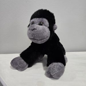 May include: A soft toy gorilla, featuring a black body and grey face, hands, and feet. The gorilla is positioned on a white surface, with its arms and legs outstretched. The toy has a fluffy texture and detailed facial features.