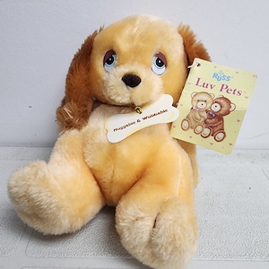 May include: A light brown plush dog with brown ears and blue eyes. The dog is sitting with its legs outstretched. The dog has a tag that says "Russ Luv Pets" and a bone-shaped tag that says "Huggable & Wubbable".