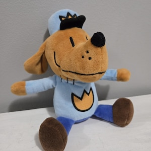 May include: A plush toy dog with a light brown face, ears, and paws, wearing a light blue outfit and hat with a black brim. The dog has a black nose and a gold emblem on its chest. The toy is sitting with its arms outstretched.