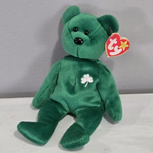 May include: A green plush bear with a white shamrock on its chest and a red heart-shaped tag. The bear has black button eyes and a black nose. A collectible stuffed animal.