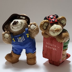Peut inclure: Deux figurines d'ours en peluche brun. L'une porte une salopette et un chapeau brun. L'autre porte un chapeau à carreaux rouge et noir et tient un traîneau rouge avec les mots "Scout's Scooter" dessus.