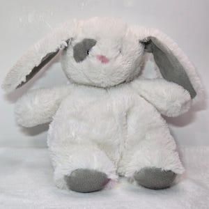 May include: A soft, white bunny toy with long ears, a grey patch over one eye, and a pink nose. The inner ears and feet are grey. The plush toy is designed to sit upright.