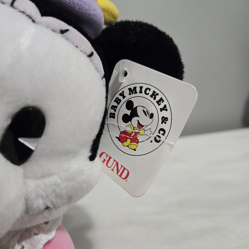 May include: A close-up of a white plush Minnie Mouse doll with black ears and a pink dress. The doll has a tag that reads "Baby Mickey & Co." and "GUND" in red.