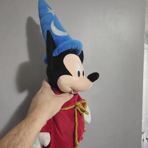 May include: A plush Mickey Mouse wearing a red robe with a gold rope tie and a blue wizard hat with a silver crescent moon and star.