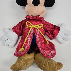 Pu&ograve; includere: Un Mickey Mouse di peluche che indossa una tunica rossa con bordi dorati e un fiocco giallo. La tunica &egrave; ricamata con note musicali. Mickey indossa stivali marroni.