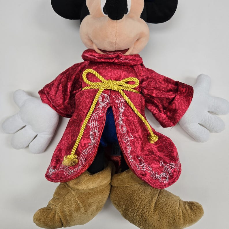 Pu&ograve; includere: Un Mickey Mouse di peluche che indossa una tunica rossa con bordi dorati e un fiocco giallo. La tunica &egrave; ricamata con note musicali. Mickey indossa stivali marroni.