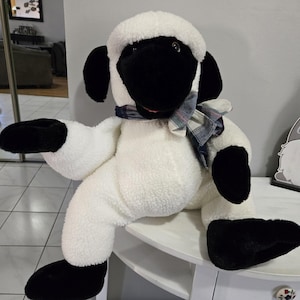 May include: A plush sheep toy with a white body, black face, ears, and feet. The sheep wears a plaid scarf around its neck. The toy is sitting on a white surface.