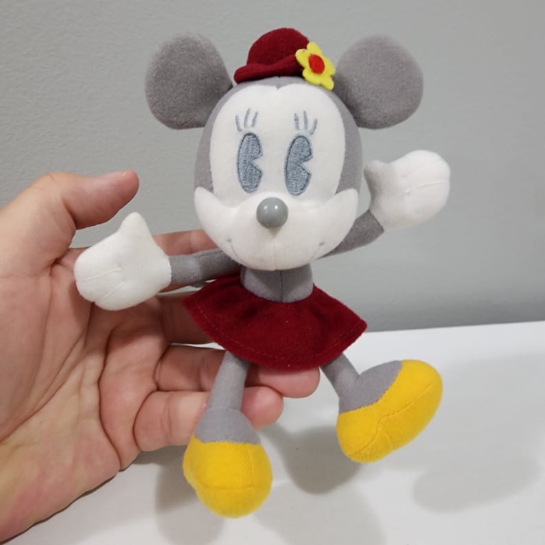May include: A gray and white plush toy mouse with red and yellow accents. The mouse is wearing a red skirt and a red hat with a yellow flower. The mouse has large, black eyes and a small, gray nose.