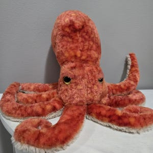 May include: A plush octopus toy in shades of orange and tan. The stuffed animal has eight long, textured tentacles and two large, yellow eyes. The octopus is sitting on a white surface.