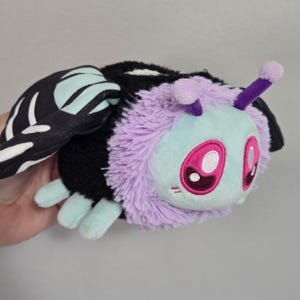 May include: A plush toy moth with a black body, lavender fur, and light blue face and feet. The moth has large pink eyes and black and white patterned wings. The antennae are purple with lavender tips.