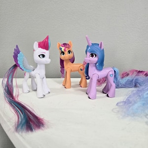 Könnte beinhalten: Drei My Little Pony Figuren, eine weiße mit einem rosa und blauen Schwanz, eine orange mit einer Regenbogenmähne und eine lila mit einem blauen und rosa Schwanz.