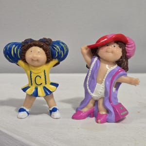 May include: Two Cabbage Patch Kids figurines. One is a cheerleader in a yellow and blue uniform with a "C" on the front. The other is wearing a purple robe, pink shoes, and a red hat. A pink purse hangs from the figurine's shoulder.