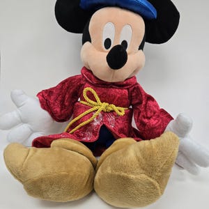 Pu&ograve; includere: Un Mickey Mouse di peluche che indossa una tunica rossa con bordi dorati e un cappello blu. &Egrave; seduto a gambe incrociate con le mani distese.