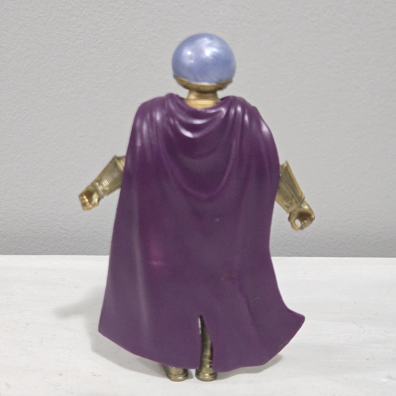 May include: A plastic action figure of a character with a purple cape, gold-colored arms, and a blue, spherical head. The figure is standing, viewed from the back, against a plain background.