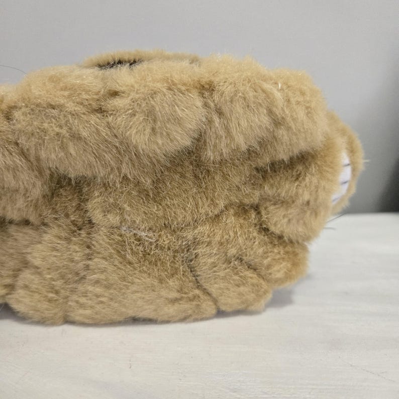 May include: Close-up of a plush toy in shades of tan and brown. The toy has a soft, furry texture and a rounded shape. A small area of black is visible on the top. The toy is resting on a white surface.