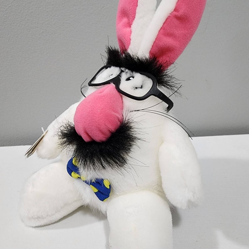 May include: A white plush bunny rabbit wearing black-rimmed glasses, a blue and yellow bow tie, and a pink nose. The bunny has a black moustache and long white fur.