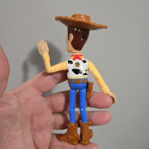 Pu&ograve; includere: Una figurina di plastica di Woody del franchise Toy Story. Woody indossa il suo cappello da cowboy marrone, il bandana rosso e i jeans blu. Sta alzando il braccio destro in segno di saluto.