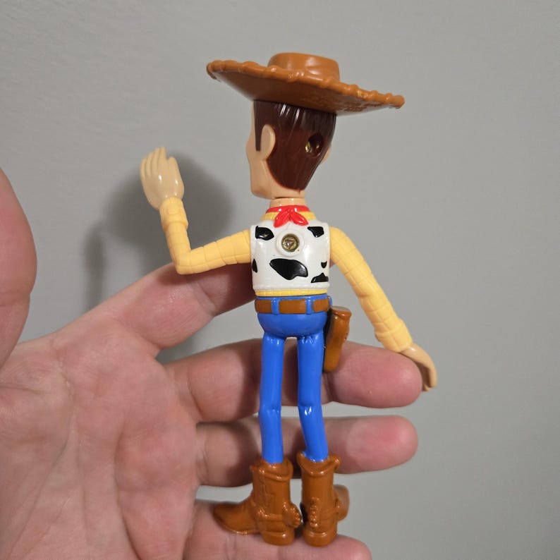 Pu&ograve; includere: Una figurina di plastica di Woody del franchise Toy Story. Woody indossa il suo cappello da cowboy marrone, il bandana rosso e i jeans blu. Sta alzando il braccio destro in segno di saluto.