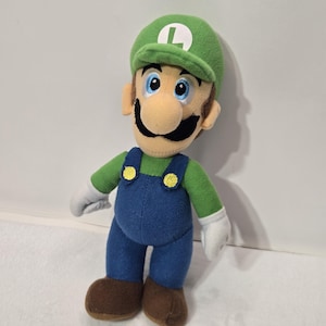 May include: A plush toy of the character Luigi, featuring a green cap with a white L emblem, a green long-sleeved shirt, blue overalls with two yellow buttons, white gloves, and brown shoes. The character has a tan face, blue eyes, and a black moustache.