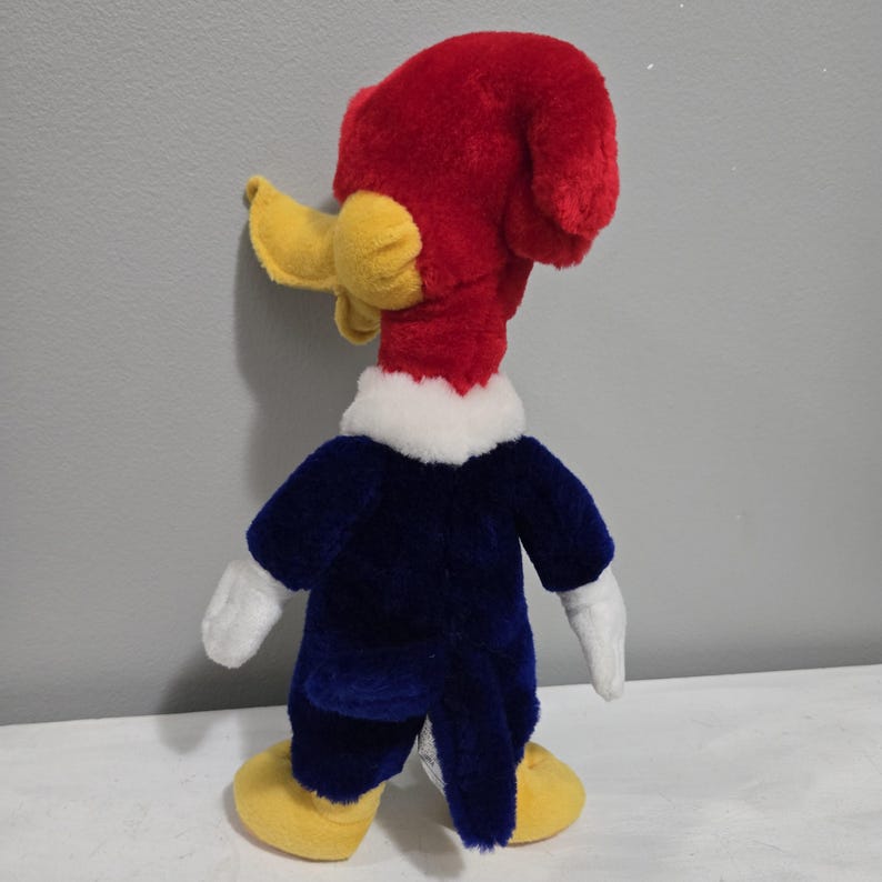 May include: A plush toy of Woody Woodpecker, a cartoon character, is shown from the back. The toy has a red head and crest, yellow beak and feet, and a blue jacket with white gloves. The toy is standing on a white surface.
