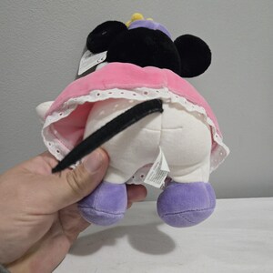 May include: A plush Minnie Mouse doll, viewed from the rear, with black ears, a pink dress with white lace trim, and purple shoes. The doll has a black tail and a purple hat with a yellow accent. A tag is attached to the doll.