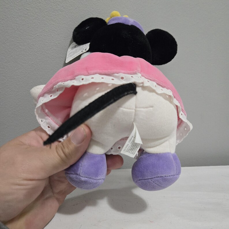 May include: A plush Minnie Mouse doll, viewed from the rear, with black ears, a pink dress with white lace trim, and purple shoes. The doll has a black tail and a purple hat with a yellow accent. A tag is attached to the doll.