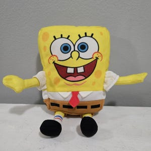 Könnte beinhalten: Ein gelbes SpongeBob Schwammkopf Plüschtier mit einem lächelnden Gesicht, blauen Augen und einer roten Krawatte. Das Spielzeug hat schwarze Schuhe, weiße Hemdärmel und eine braune Hose. Die Arme sind ausgestreckt.