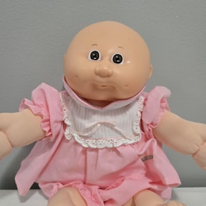 Könnte beinhalten: Vintage Cabbage Patch Kid Puppe mit kahlem Kopf und weichem, rundem Gesicht. Die Puppe trägt ein rosa Kleid mit Rüschenärmeln und ein weißes Lätzchen mit Spitzenbesatz. Die Puppenaugen sind braun und sie hat einen kleinen, lächelnden Mund.