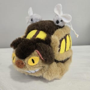 May include: A plush toy resembling a character from an animated film. The toy is primarily tan and brown, with yellow eyes and accents. Two small, gray and white mouse figures sit on top of the toy.