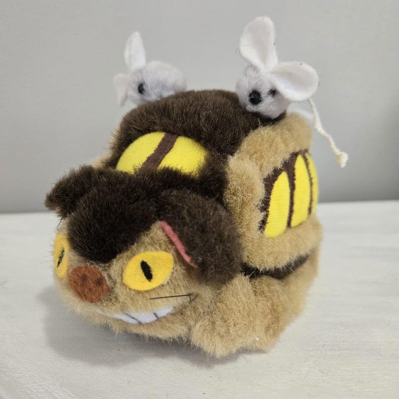 May include: A plush toy resembling a character from an animated film. The toy is primarily tan and brown, with yellow eyes and accents. Two small, gray and white mouse figures sit on top of the toy.
