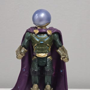 May include: A Mysterio action figure with a metallic blue helmet and a purple cape. The figure has a green and gold armored suit with intricate details. The cape is a deep purple color, and the figure is standing upright.