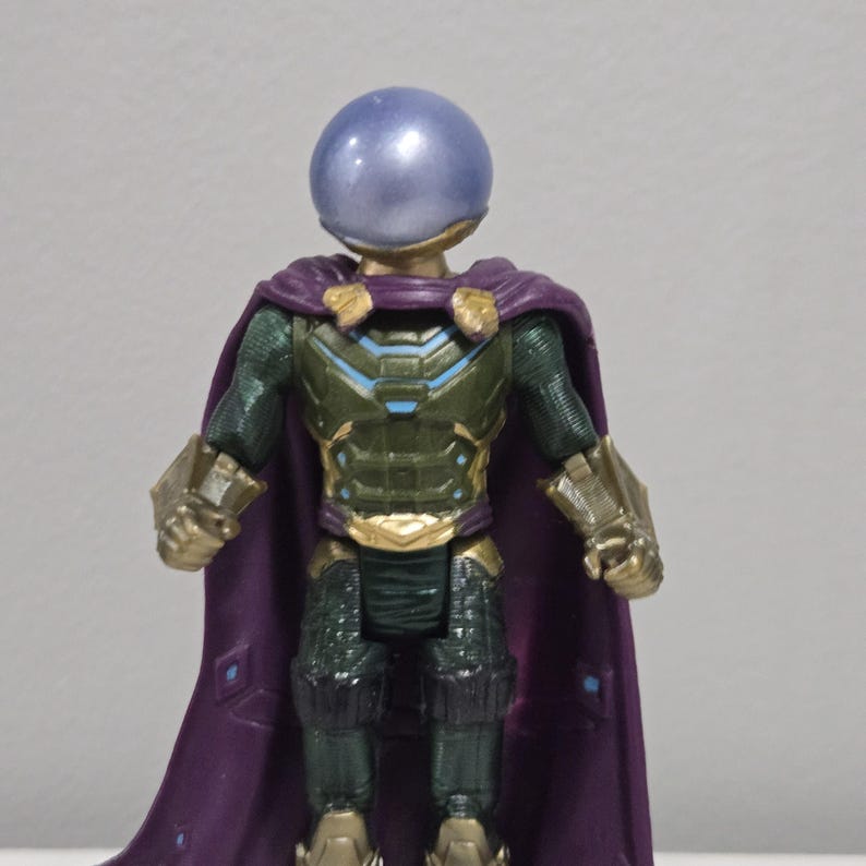 May include: A Mysterio action figure with a metallic blue helmet and a purple cape. The figure has a green and gold armored suit with intricate details. The cape is a deep purple color, and the figure is standing upright.