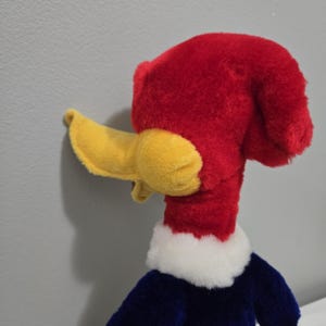 May include: A plush toy of Woody Woodpecker, featuring a bright red head and a large yellow beak. The toy has a white collar, a blue body, and is made of soft, plush material. The iconic cartoon character is a classic.