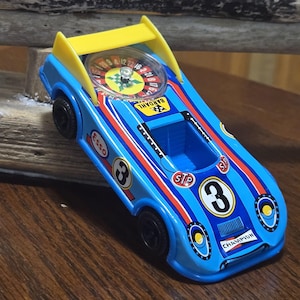 以下が含まれることがあります： 黄色いリアスポイラー、黒いホイール、車体に組み込まれたルーレットホイールを備えた青いおもちゃのレーシングカー。車体には数字の3、Champion、Bardahl、Essoの文字、ボンネットには赤とオレンジのレーシングストライプが施されています。