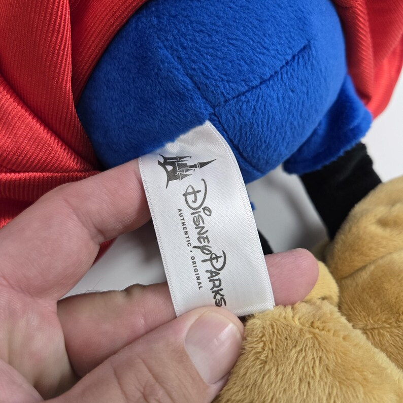 Pu&ograve; includere: Un'etichetta bianca con il testo "Disney Parks Authentic. Original" e un logo di castello nero. L'etichetta &egrave; attaccata a un giocattolo di peluche.