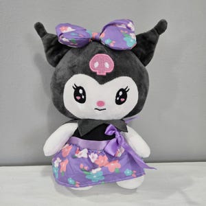 May include: A plush toy of Kuromi, a black and white character from the Sanrio franchise. Kuromi is wearing a purple dress with pink and blue flowers and a purple bow. She has a pink skull on her forehead and big, black eyes.